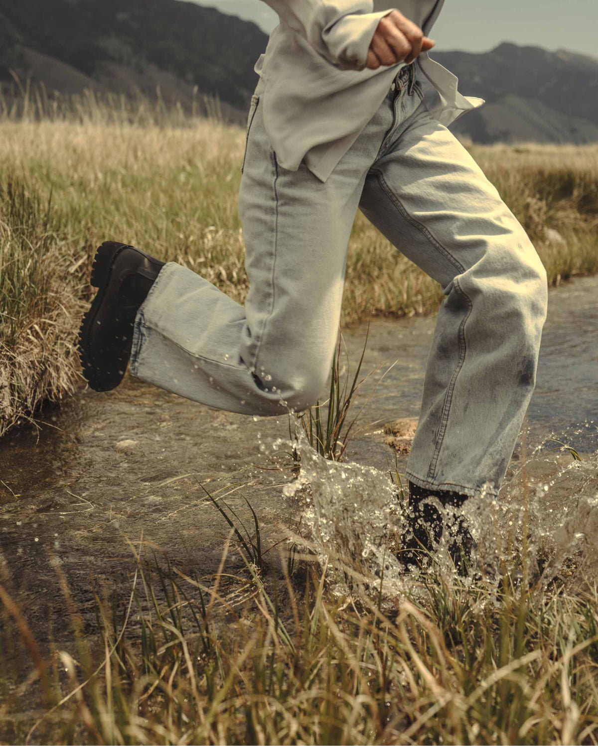 A person running through shallow water in an outdoor grassy landscape, wearing light-wash wide-leg jeans, a light gray top, and black boots.