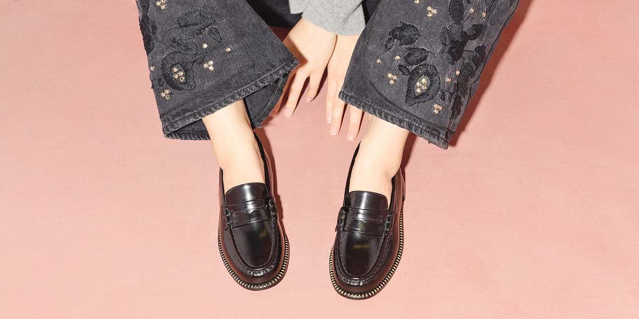 Overhead view of a model seated on a blush pink background wearing the FREDA SALVADOR ELBA Penny Loafer in Black Calf, styled with black embroidered denim and a grey sweater.