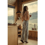 A woman stands by a large window in a rustic wooden cabin, looking out at a mountain and lake landscape. She is wearing a light beige ruffled blouse, light-wash wide-leg jeans, and brown boots.