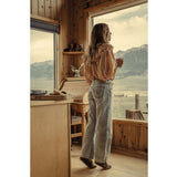 A woman stands by a large window in a rustic wooden cabin, looking out at a mountain and lake landscape. She is wearing a light beige ruffled blouse, light-wash wide-leg jeans, and brown boots.