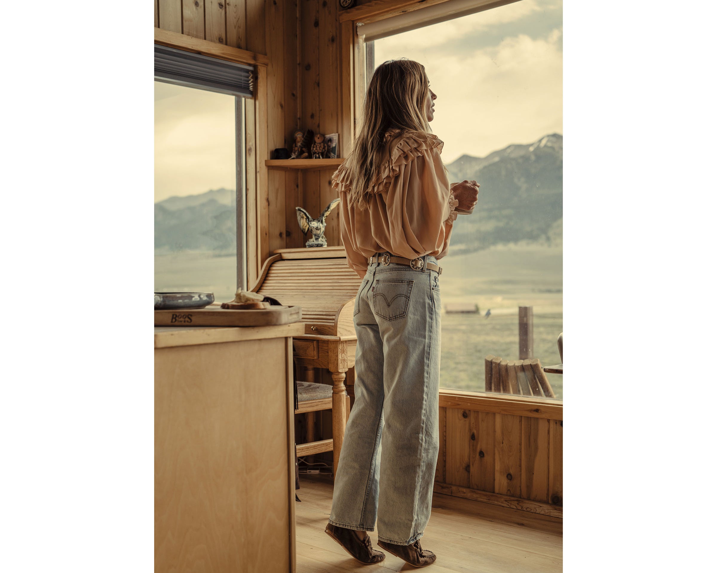 A woman stands by a large window in a rustic wooden cabin, looking out at a mountain and lake landscape. She is wearing a light beige ruffled blouse, light-wash wide-leg jeans, and brown boots.
