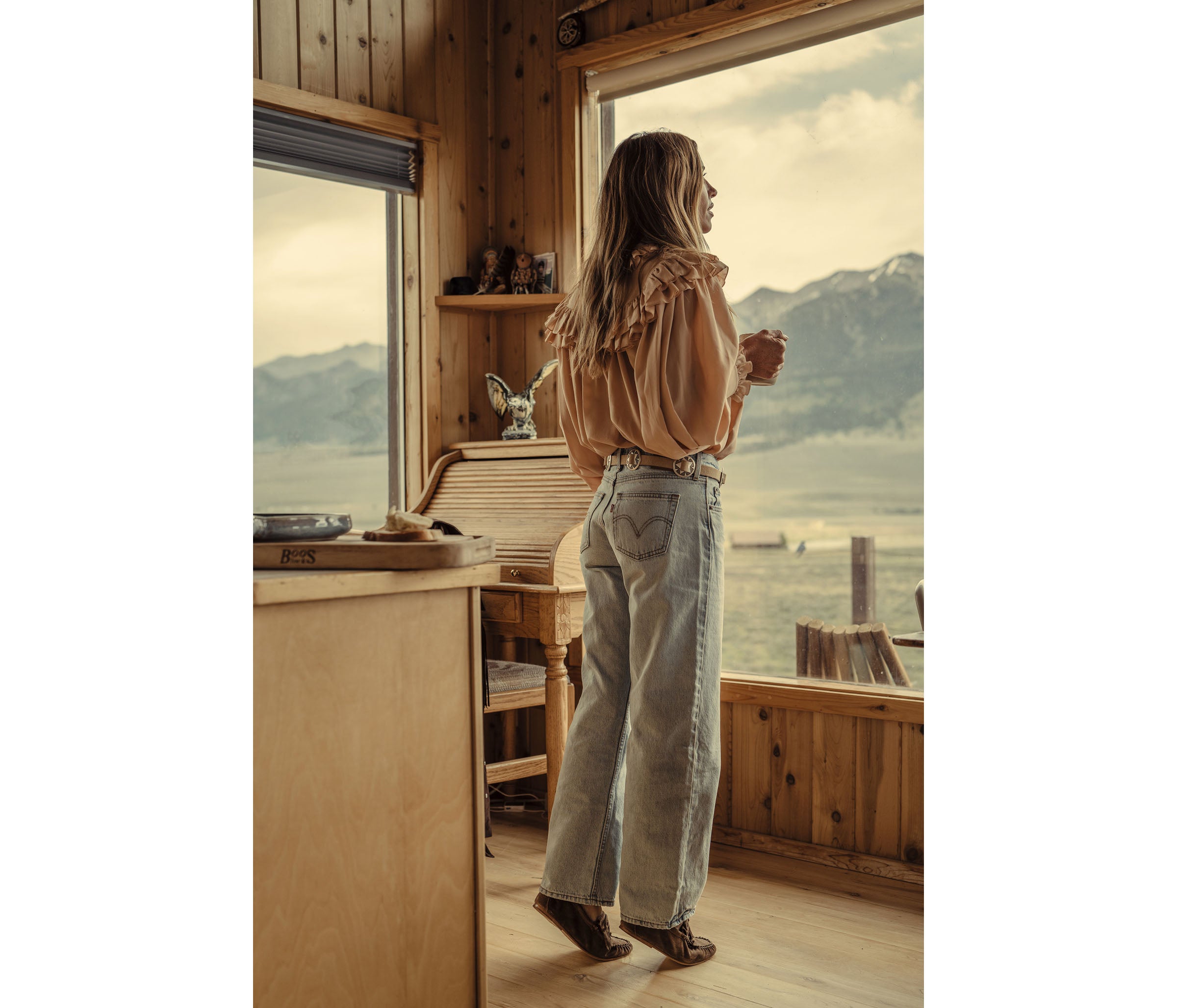 A woman stands by a large window in a rustic wooden cabin, looking out at a mountain and lake landscape. She is wearing a light beige ruffled blouse, light-wash wide-leg jeans, and brown boots.